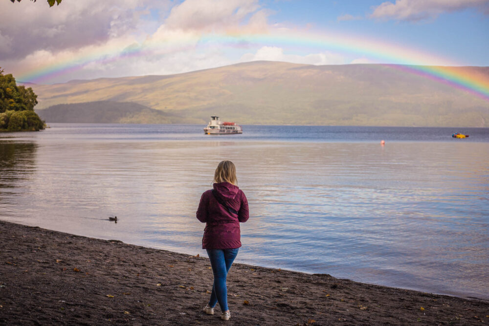 Woman standing by Loch Lomond with a rainbow arching overhead