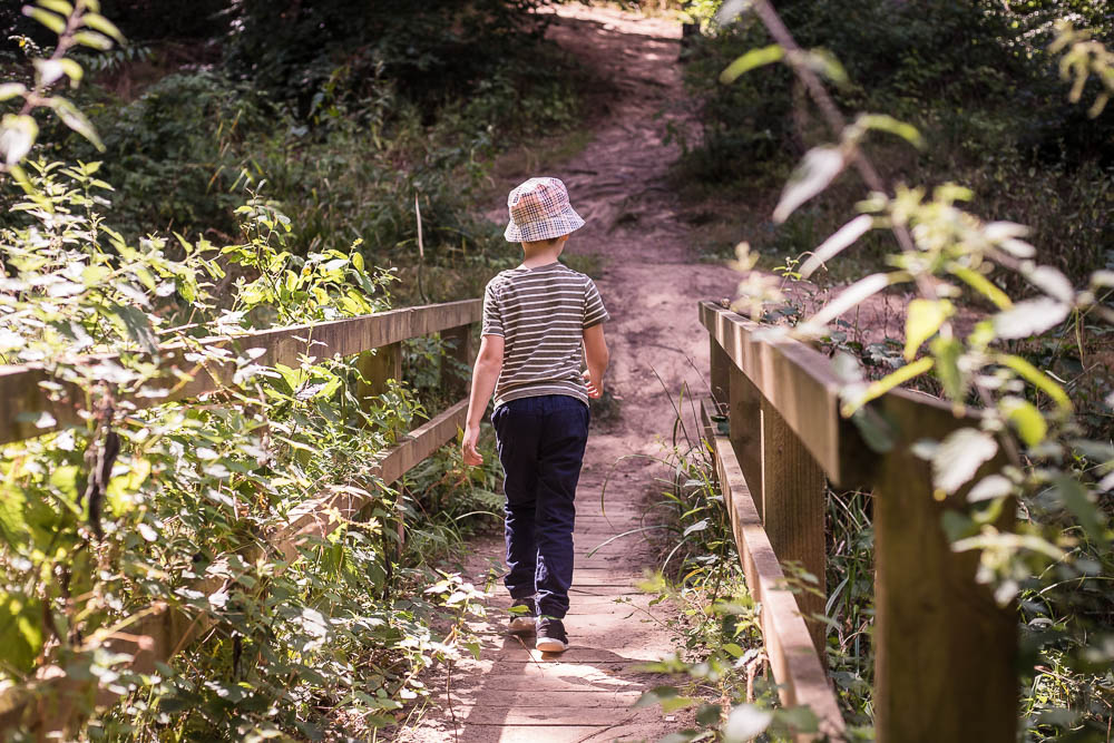 Child walking away from the camera over a small wooden bridge in a woodland area, wearing a bucket hat.