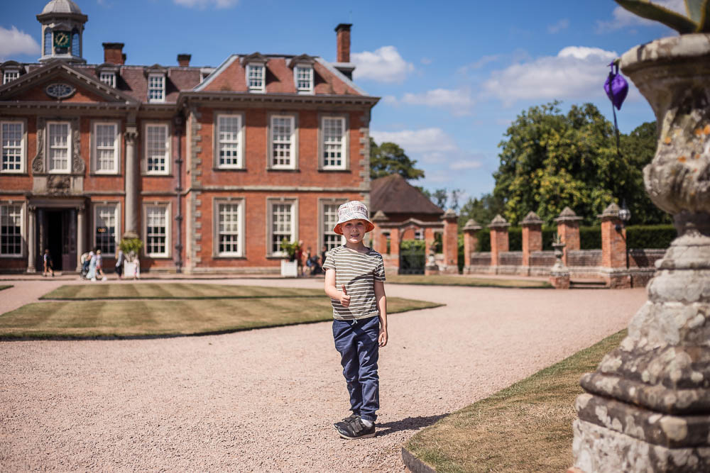 Pickle stood outside Hanbury Hall wearing a bucket hat and holding a thumbs up at the National Trust property